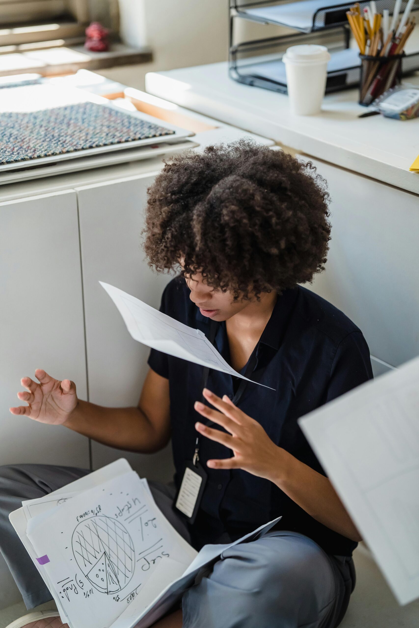 A frustrated professional sits on the floor throwing papers amid office work chaos.